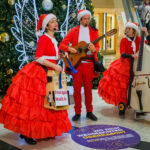 drei Musiker*innen in weihnachtlichen Kostümen mit Instrumten im Einkaufszentrum singen für die Kunden der Shopping Mall