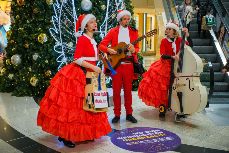 drei Musiker*innen in weihnachtlichen Kostümen mit Instrumten im Einkaufszentrum singen für die Kunden der Shopping Mall
